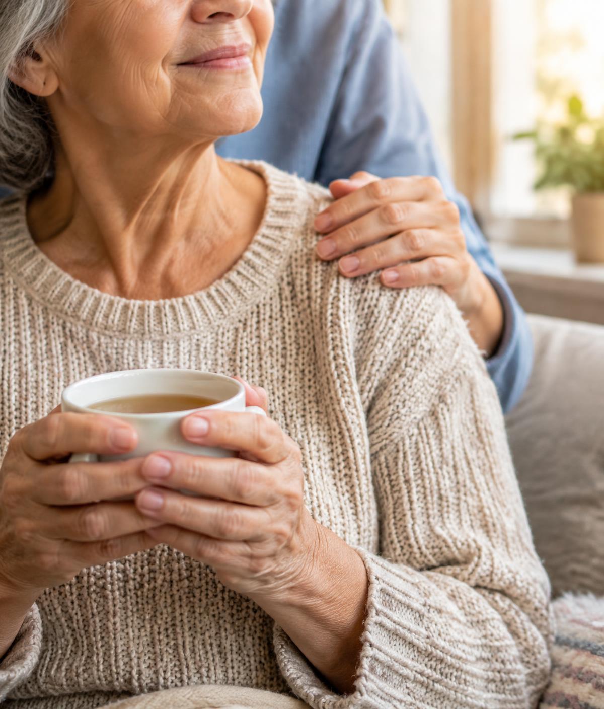An older woman holding a cup of tea as a caregiver rests a hand on her shoulder
